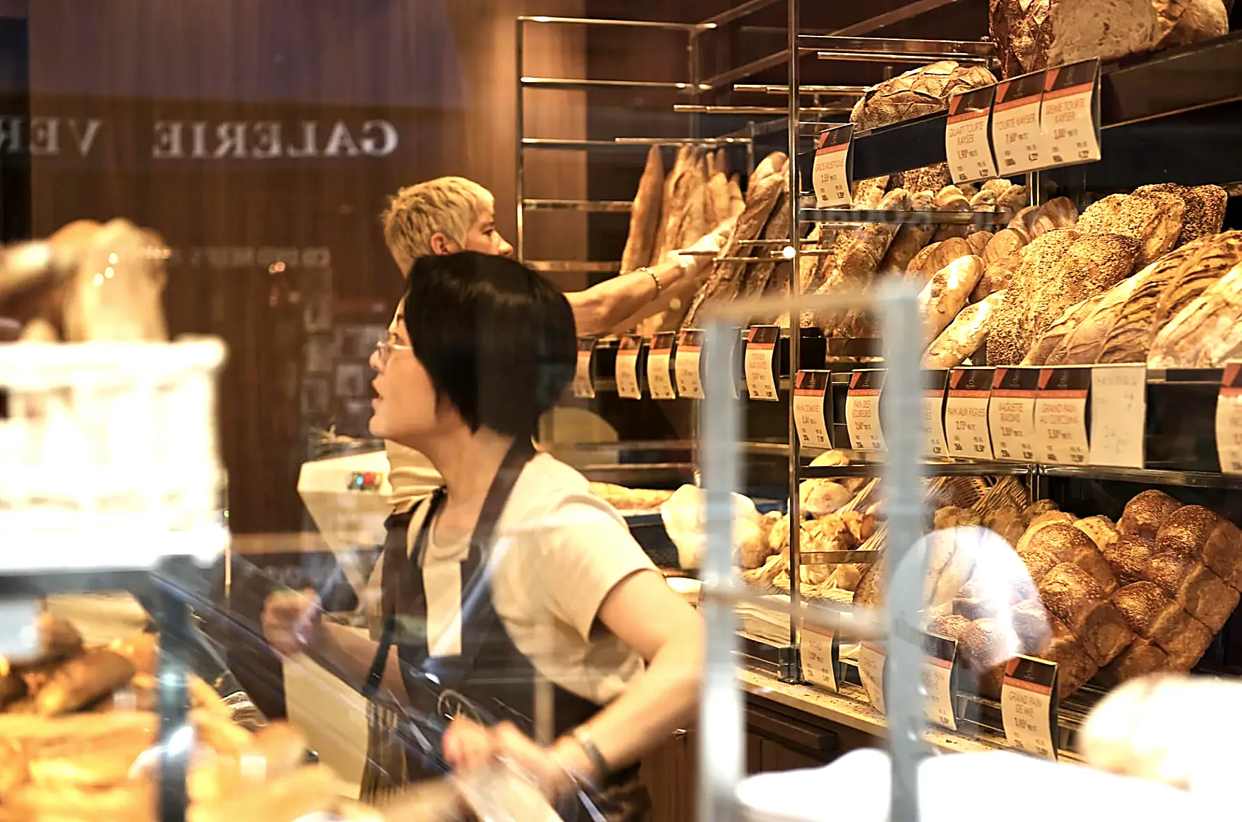 A baker serving at a bread counter stacked with loaves